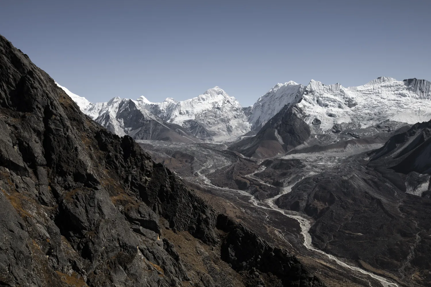 Looking past a steep, rocky dark mountain slope in the foreground, a wide glacier valley unfolds below, leading toward a dramatic backdrop of massive, snow-covered Himalayan peaks under a pale blue sky.