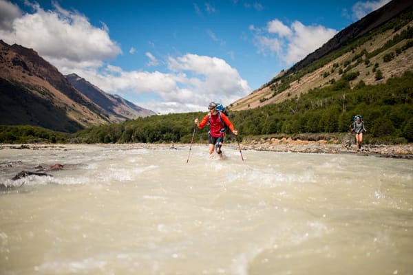 A hiker crosses a fast-flowing glacial river with mountains in the background.