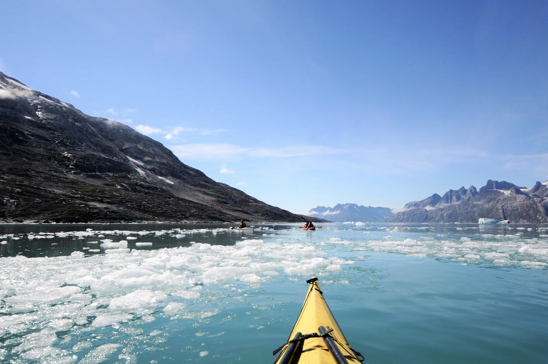 View from a yellow kayak paddling through light blue water filled with drift ice toward two other kayaks, surrounded by massive mountains under a clear blue sky.