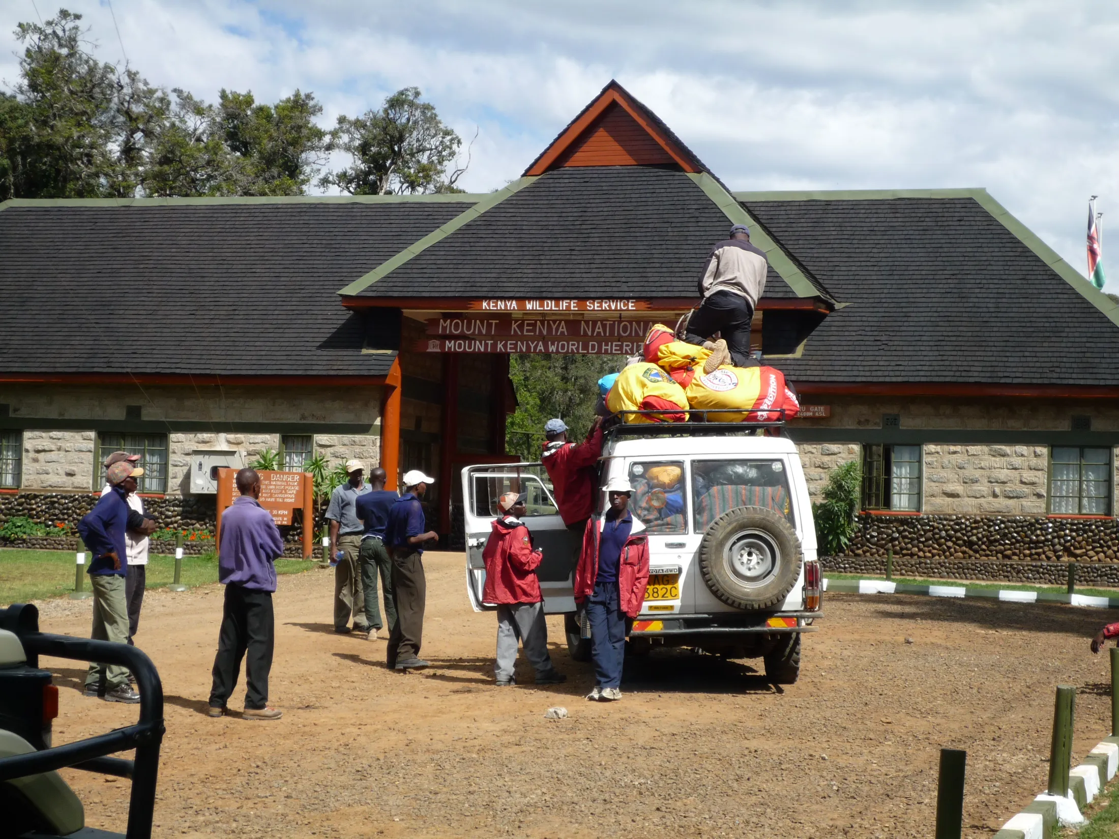 A group of people unloads large gear bags from the roof of a white off-road vehicle parked in front of the Kenya Wildlife Service entrance gate to Mount Kenya National Park.