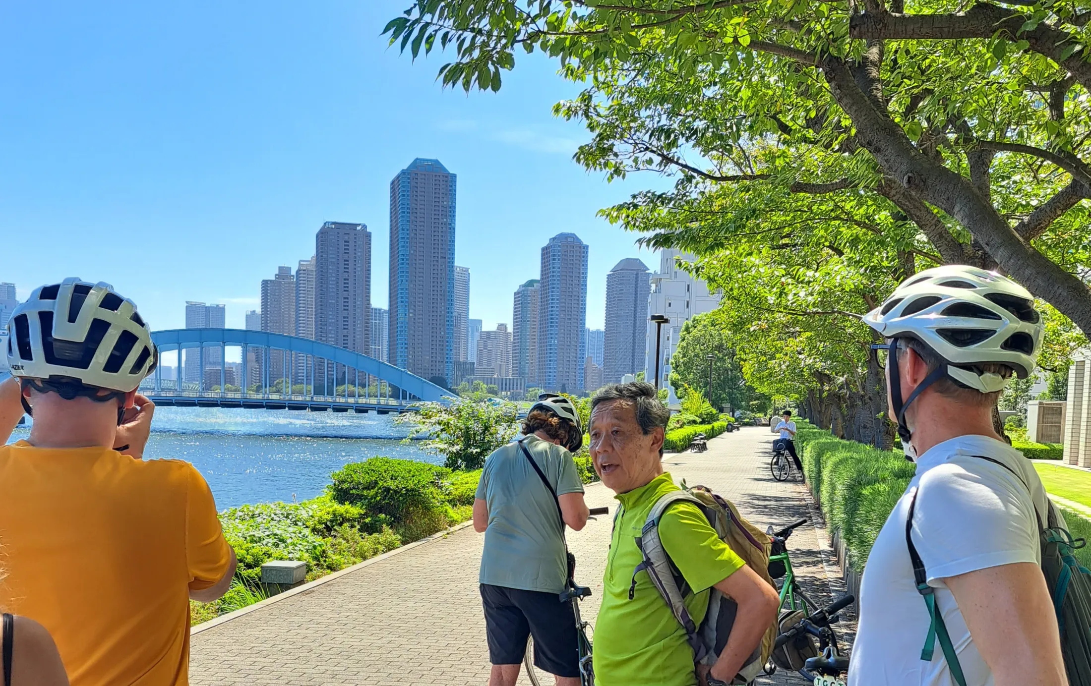 A group of cyclists with helmets pauses along a riverside paved pathway to talk, with a blue arch bridge and modern city high-rises visible across the water.