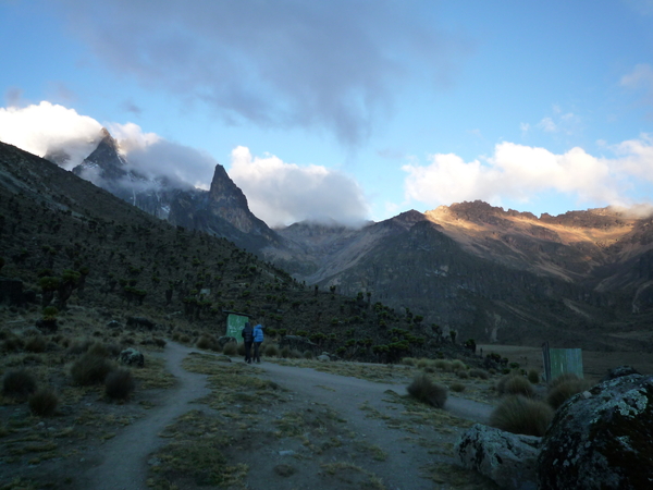 Two hikers walk along a dirt path in a vast mountain valley on Mount Kenya, with dramatic rock peaks partially covered by low clouds in the background.