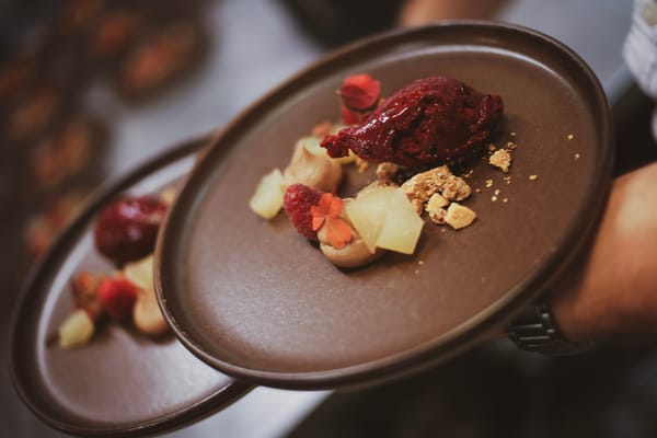 A person holds two brown plates topped with a gourmet dessert including a scoop of dark berry sorbet, crumble, and fruit pieces.