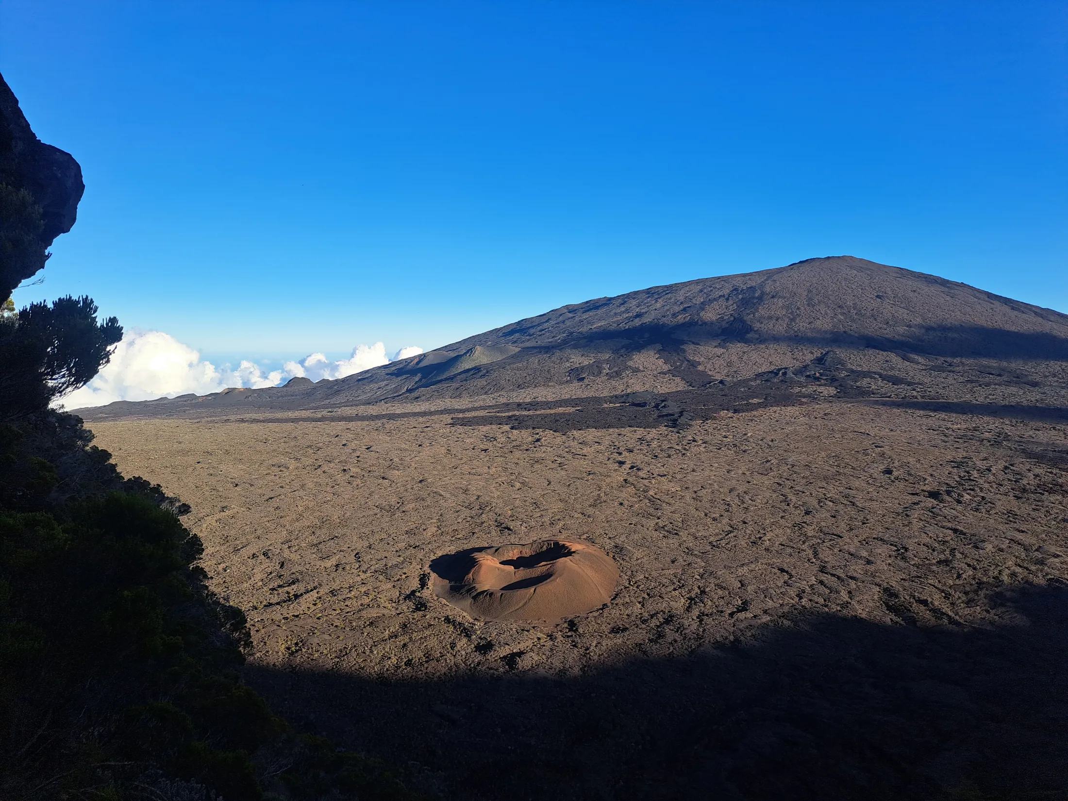 A vast, barren volcanic landscape featuring a small reddish crater in the foreground and a large mountain peak under a clear blue sky.