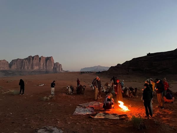 A group of people and camels gathered around a campfire in a vast desert landscape at dusk, with dramatic mountain formations in the background.