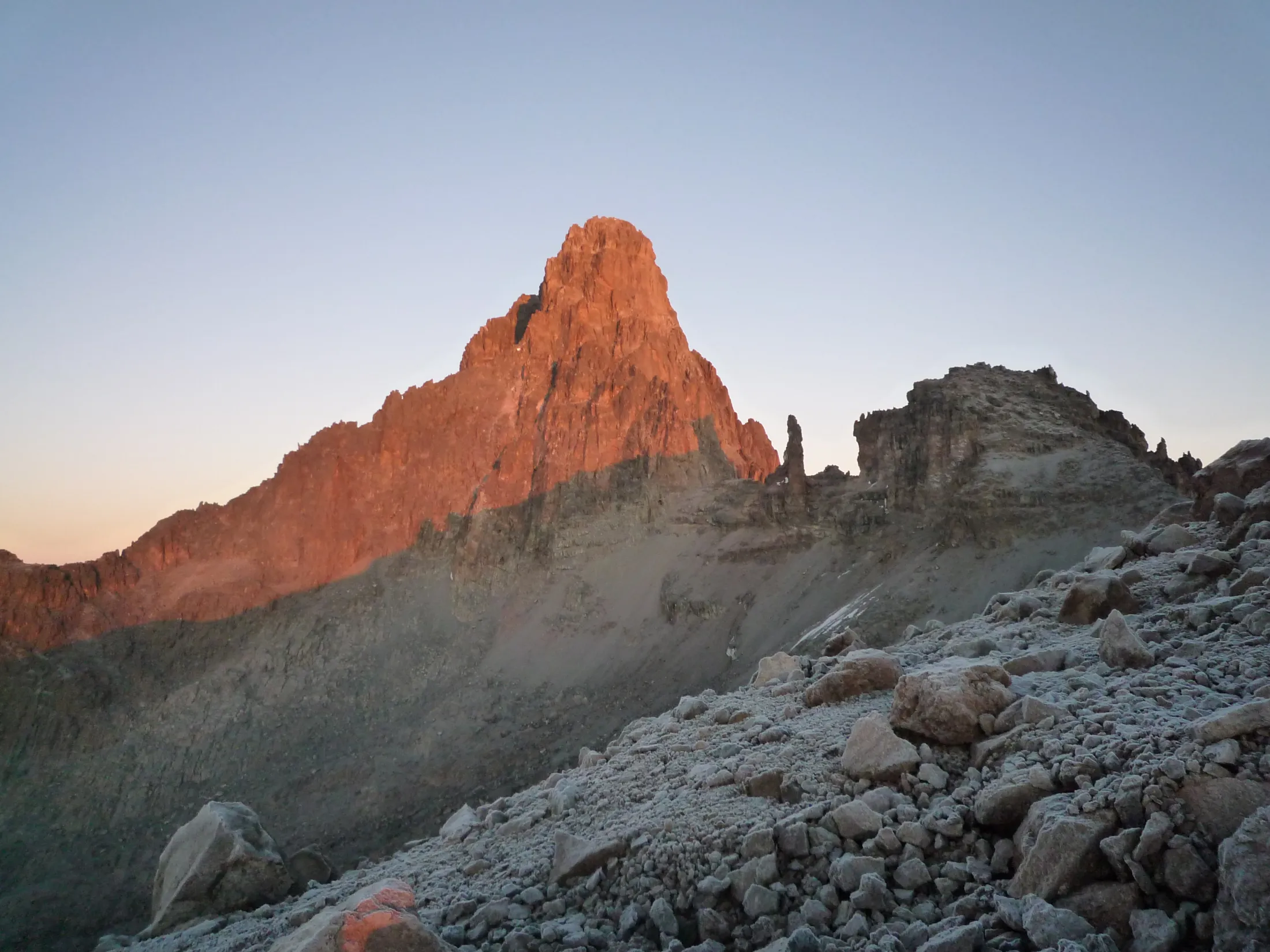 The towering rocky summit of Mount Kenya glows with a warm orange light from the rising sun, viewed from a cold, scree-covered alpine slope at dawn.