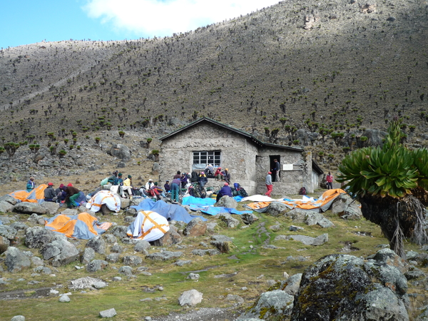 A group of hikers rests outside a stone mountain hut surrounded by tents and gear on the rocky, alpine slopes of Mount Kenya.
