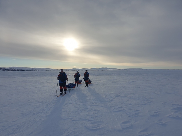 Three people backcountry cross-country skiing across a vast snow-covered landscape beneath a bright, cloudy sky.
