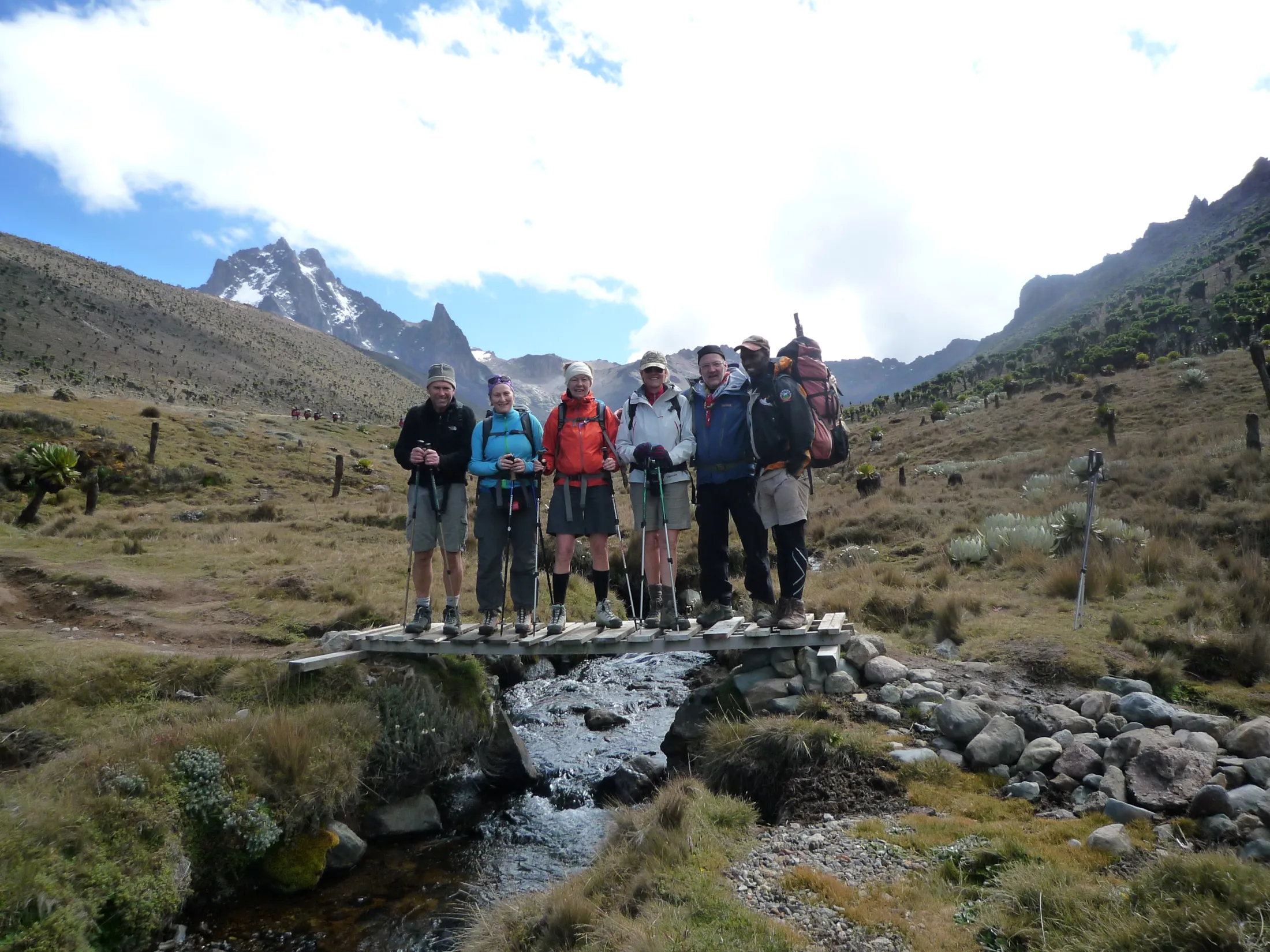 A group of six hikers stands smiling on a small wooden footbridge over a rocky mountain stream, with the craggy peaks of Mount Kenya rising in the background.