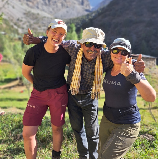 A smiling man and two female hikers stand with their arms around each other, giving a thumbs-up gesture in a sunny outdoor setting.
