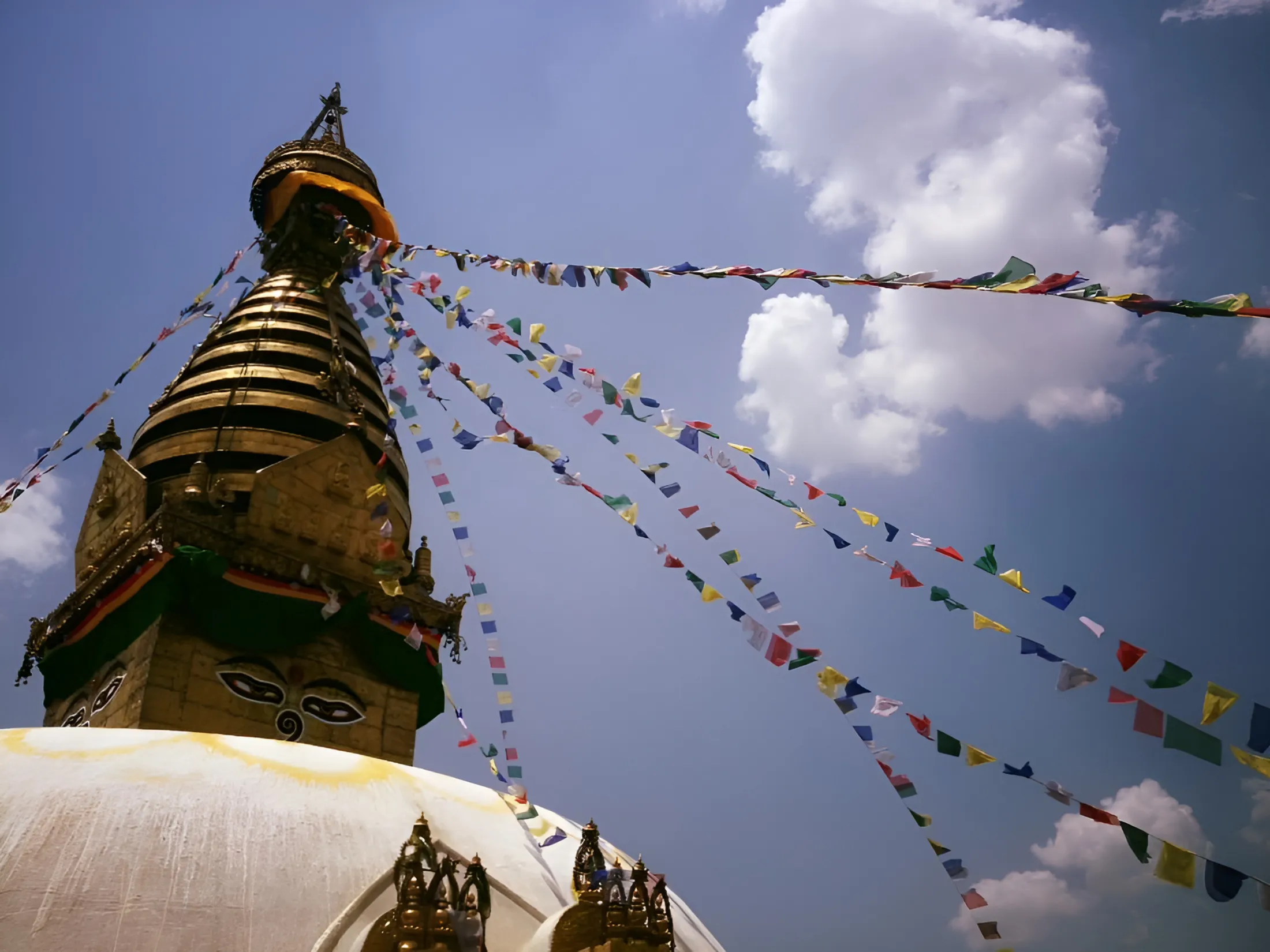 A low-angle shot of a golden Buddhist stupa adorned with the eyes of Buddha, with colorful prayer flags stretching out against a bright blue sky with scattered clouds.