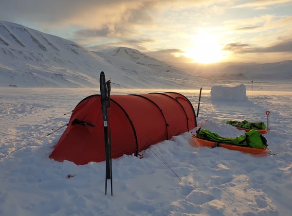 A red tent pitched in the snow with skis planted upright outside, set against a snow-covered mountain landscape during a bright sunset.