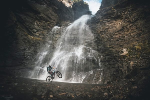A mountain biker performing a stunt in front of a majestic waterfall cascading down a rocky cliff.