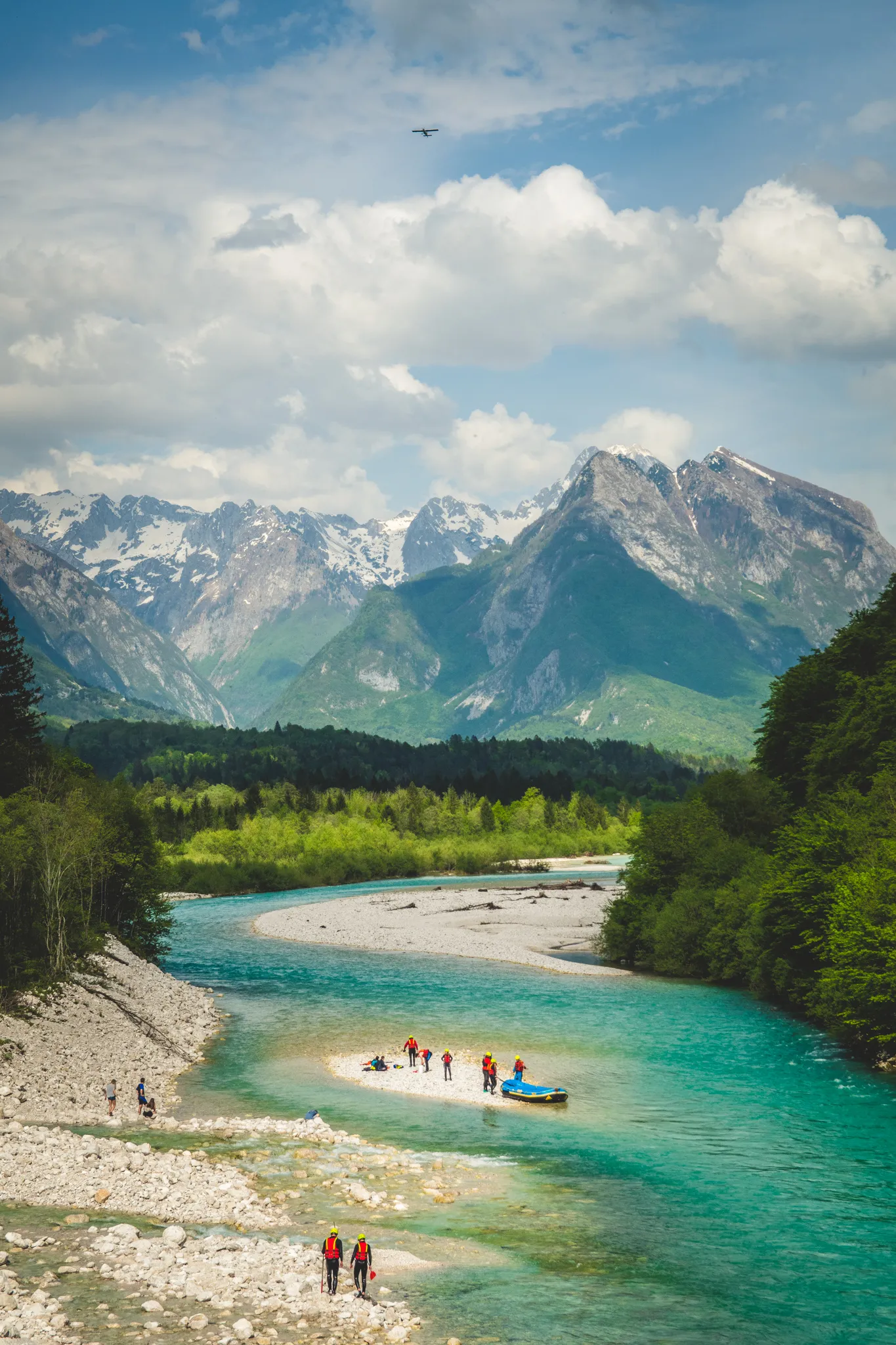 A group of rafters gathers on a white gravel sandbar in a vibrant turquoise river, with lush green forests and snow-capped mountain peaks rising under a cloudy sky.