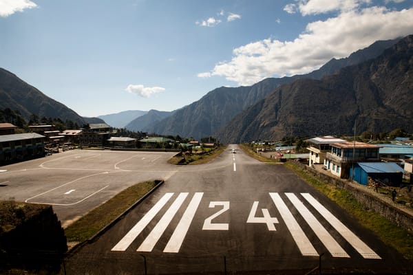 Looking down a short, sloped asphalt airport runway marked with the number 24, nestled in a mountain valley village with a small airplane visible in the distance under a partly cloudy sky.