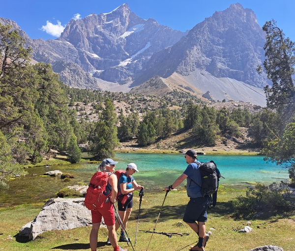 Three hikers with backpacks stand on a grassy shore, talking near a vibrant turquoise mountain lake surrounded by trees and towering, rugged peaks.