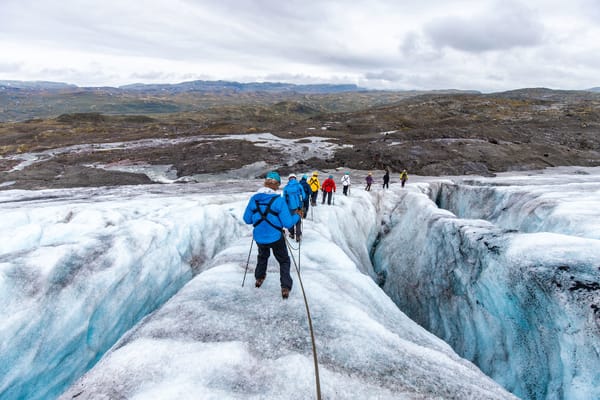 A group of roped hikers wearing helmets walks along a narrow, icy ridge of a glacier next to a deep crevasse.