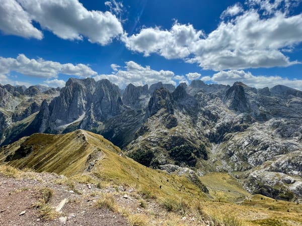 Rugged mountain range under a blue sky with scattered white clouds, viewed from a grassy ridge.