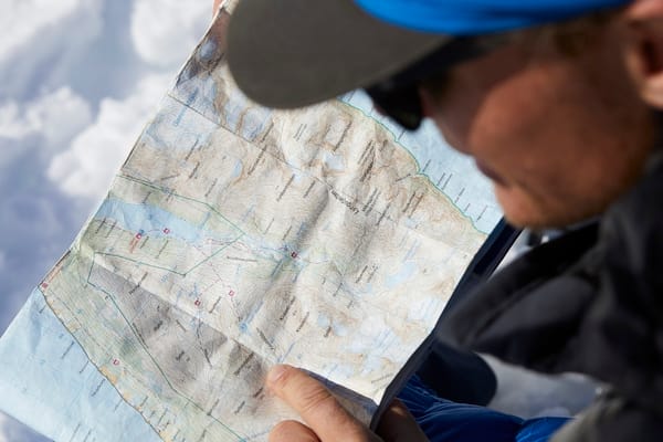 A person wearing a cap and jacket looks closely at a detailed topographic map outdoors.