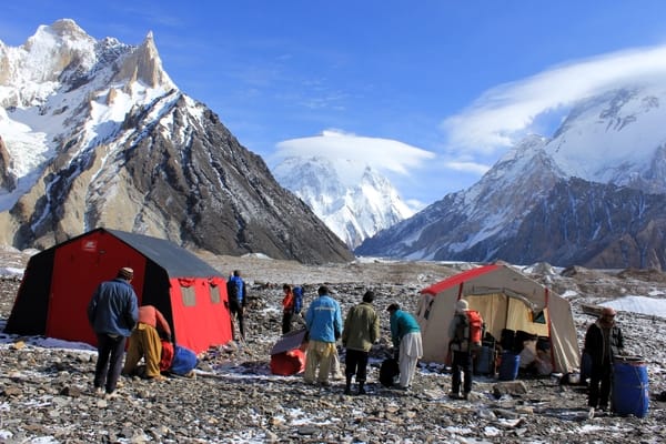 People gather outside large expedition tents set up on a rocky mountain terrain, with massive snow-capped peaks rising in the background beneath a clear sky.