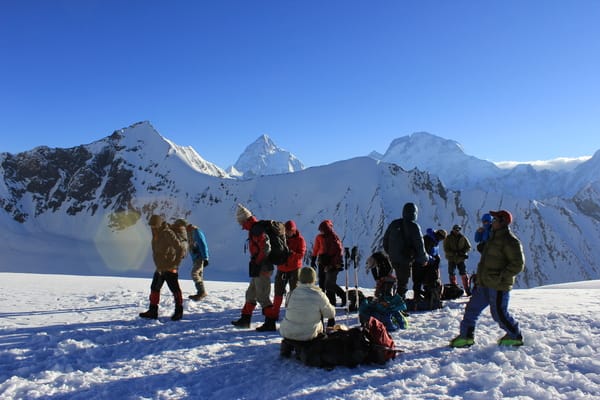 A group of mountaineers wearing warm winter jackets gear up on a snowy peak under a clear blue sky, with high mountain ranges in the background.