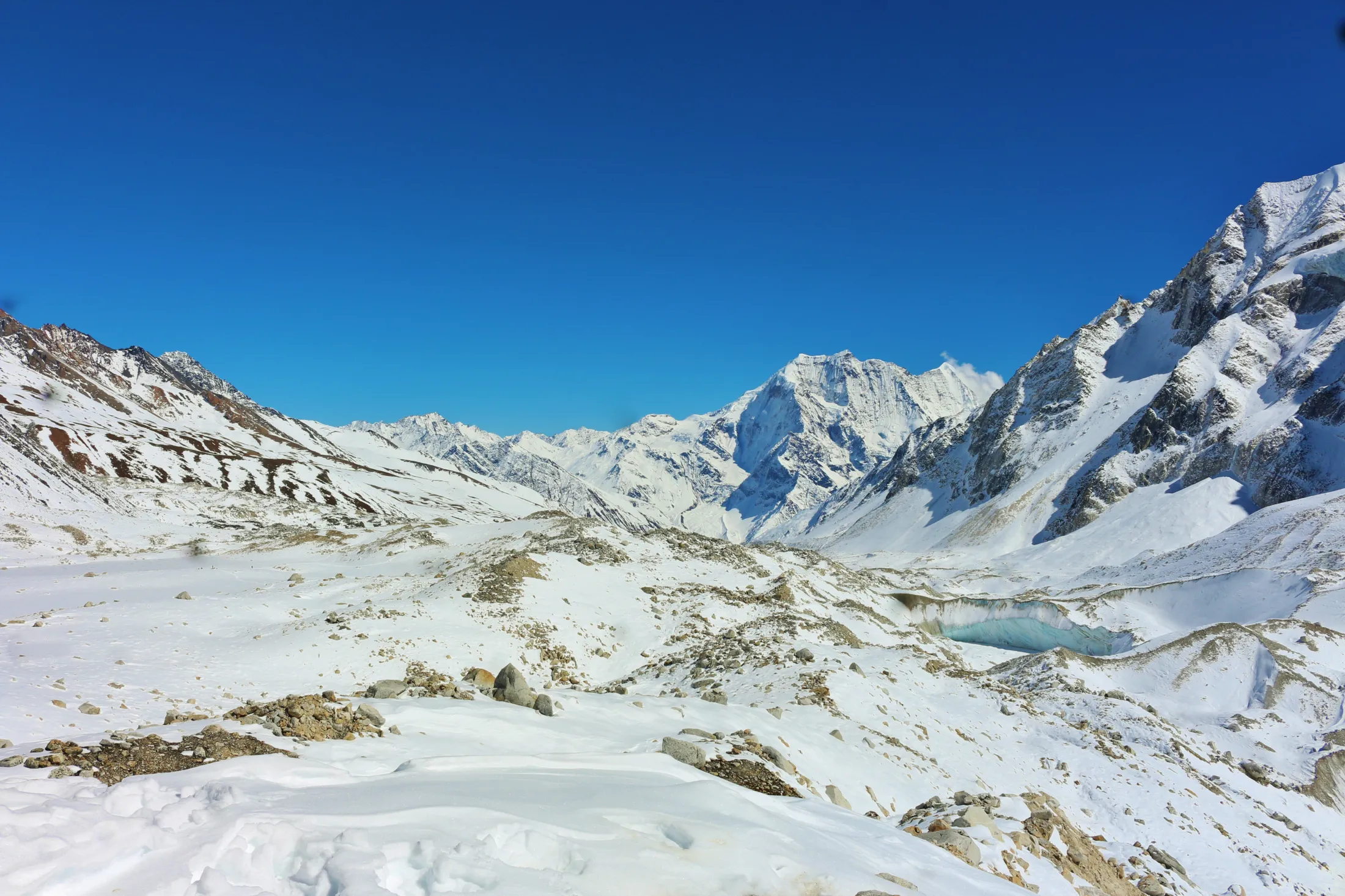 A vast, snow-covered mountain landscape opens up under a clear blue sky, featuring rocky ridges and a glimpse of a frozen, light-blue glacial lake.