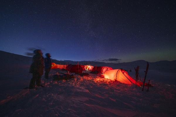 Glowing red tents pitched in the snow under a clear starry night sky, with two figures looking up at the stars.