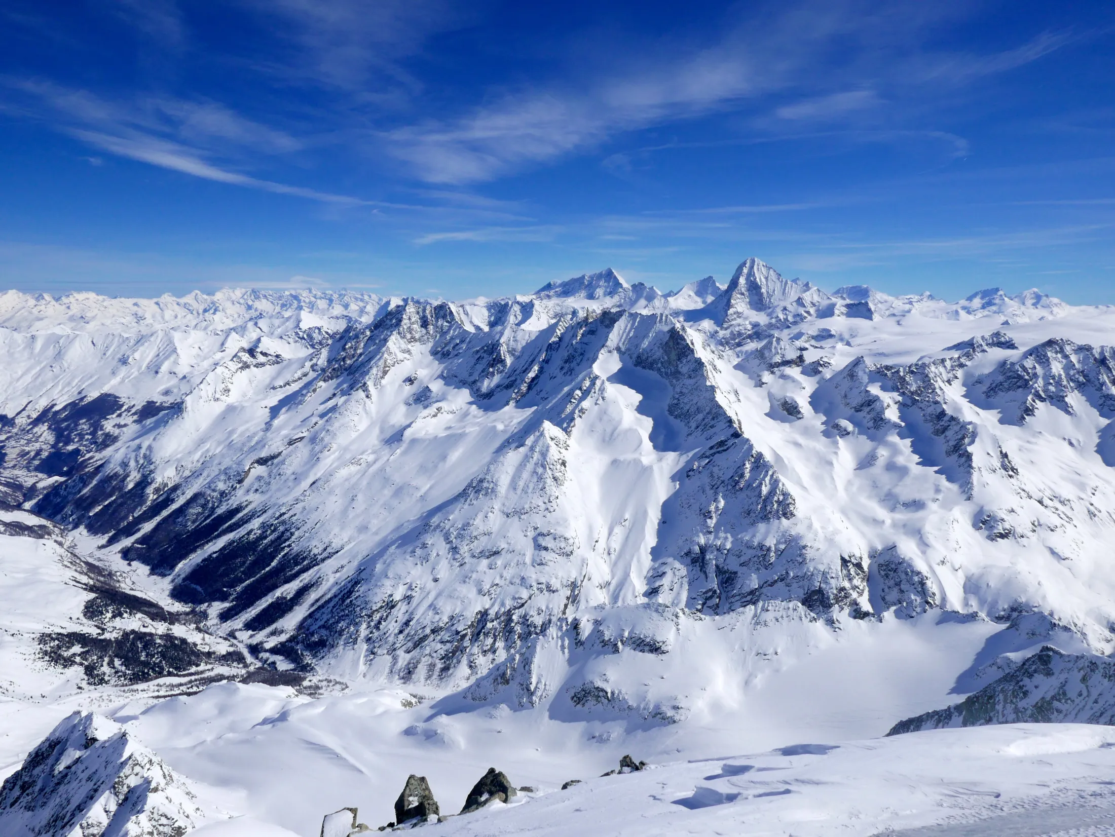 A breathtaking panoramic view shows vast, snow-covered mountain ranges and deep valleys under a clear blue sky with light, wispy clouds.