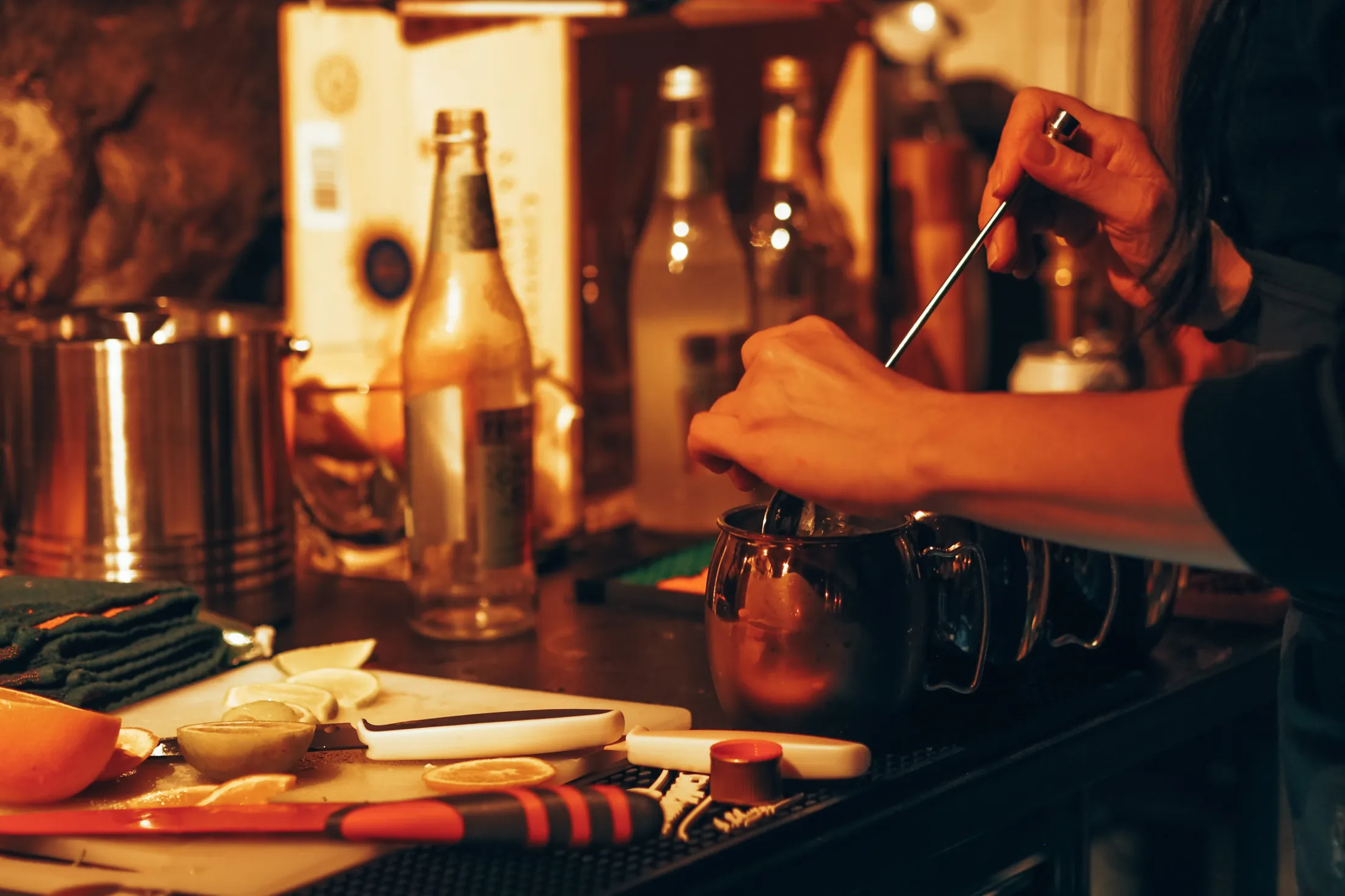 A bartender stirs a drink in a metal mug on a bar counter filled with citrus fruits, knives, and bottles under warm lighting.