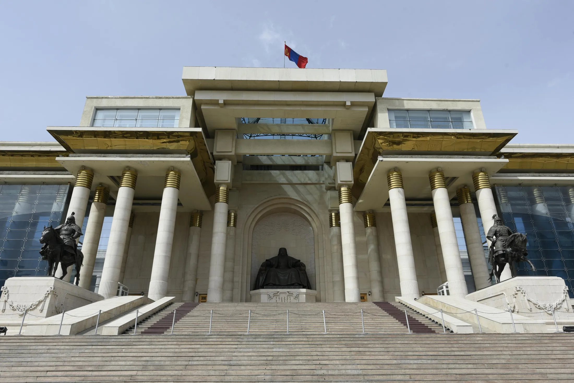 The grand facade of the Government Palace in Ulaanbaatar features a large bronze statue of Genghis Khan seated at the top of a wide stone staircase under the Mongolian flag.