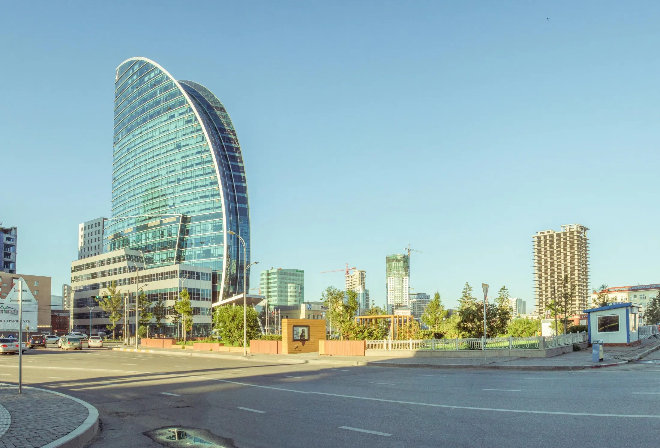 The distinctive glass facade of the sail-shaped Blue Sky Tower rises above a city street and a small green plaza under a clear, bright sky.