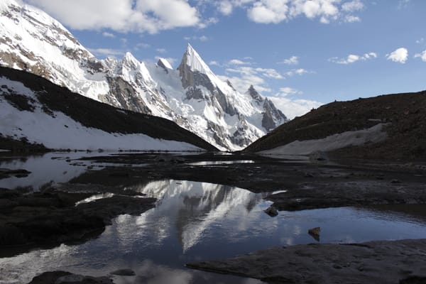 A sharp, snow-covered mountain peak is reflected in a calm pool of water set among rocky ground and snowy slopes under a partly cloudy sky.