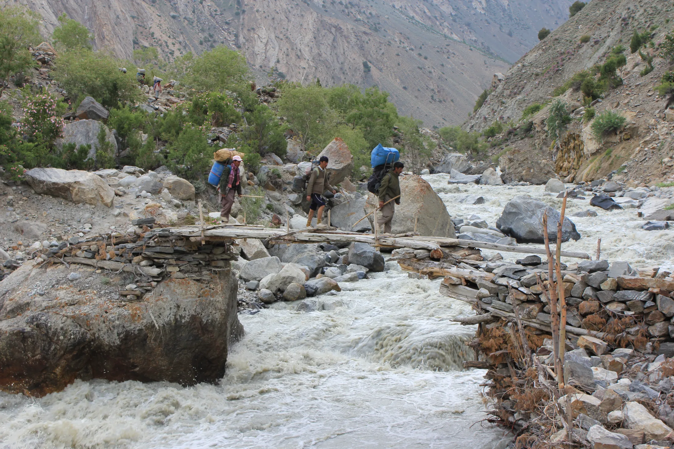 Porters carrying heavy loads cross a rustic wooden footbridge over a rushing mountain river in a rocky valley.