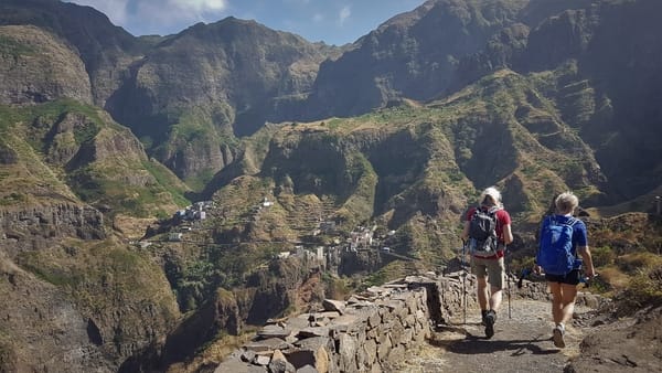 A hiker with a backpack and trekking poles walks along a stone-lined trail overlooking a mountain village nestled in a dramatic, terraced green valley.