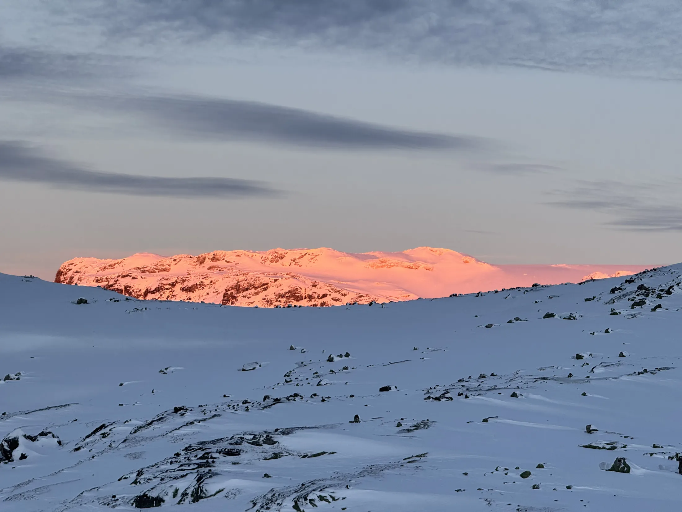 Snowy landscape with a mountain peak illuminated in pink and orange by the setting sun under a cloudy sky.