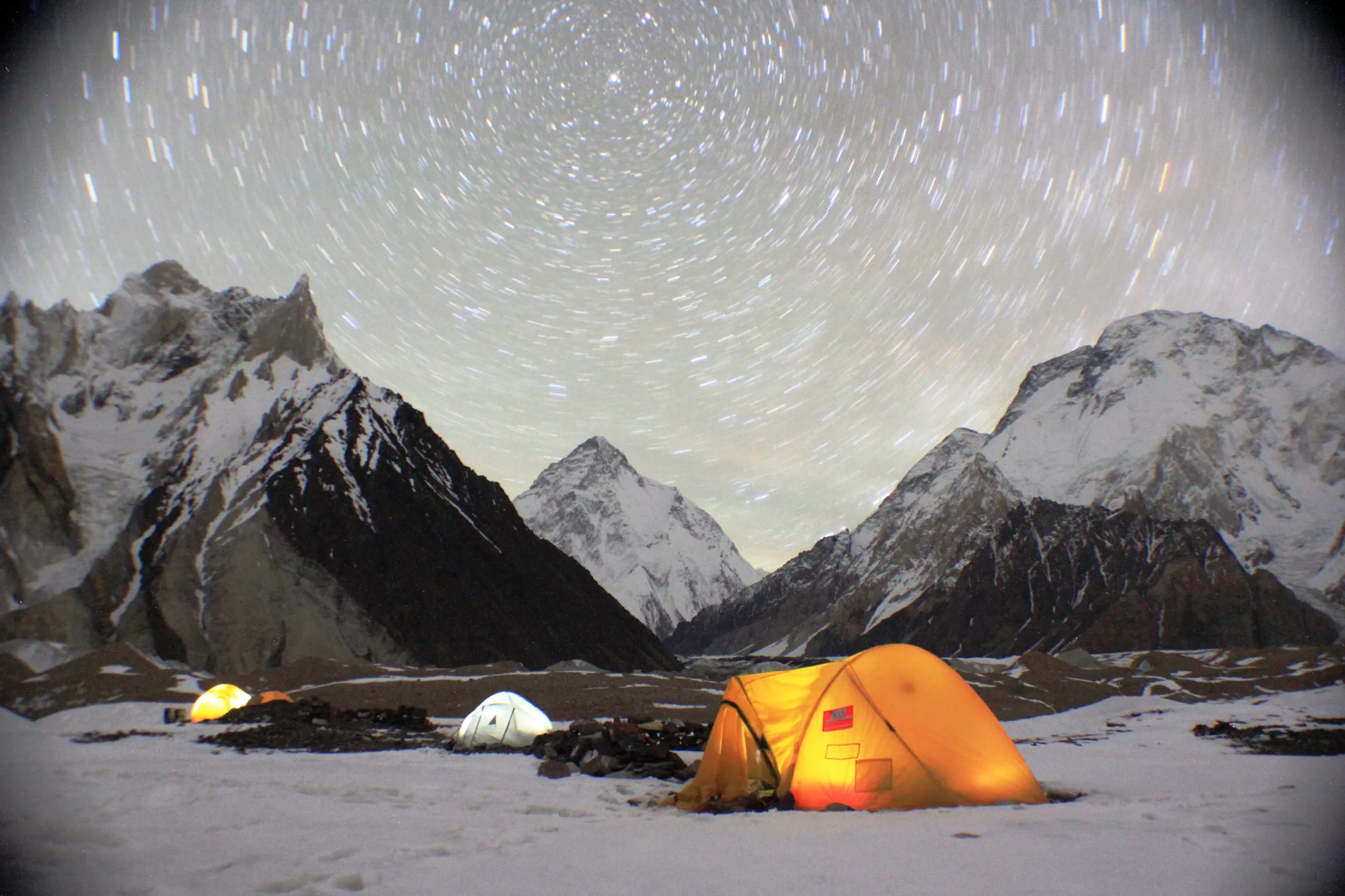 Glowing tents illuminate a snow-covered campsite at night beneath a swirling star trail sky framed by dark, snow-dusted mountains.