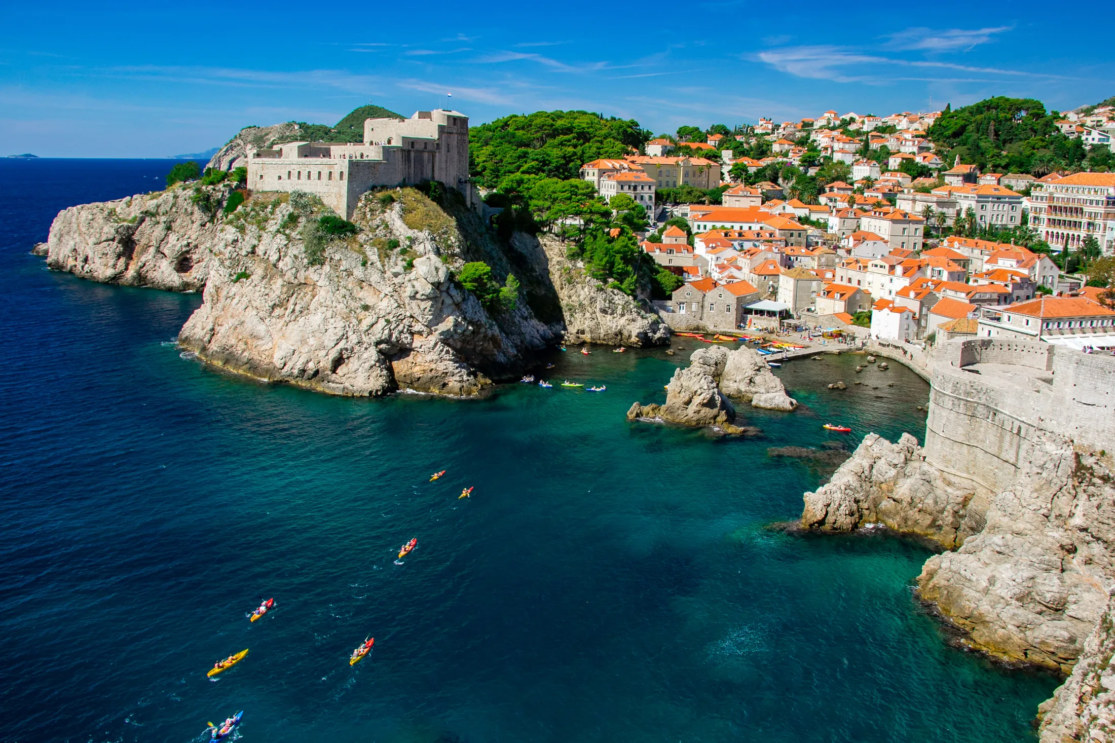 View of Dubrovnik's coastline with white houses, a historic castle, and colorful kayaks in the bay.
