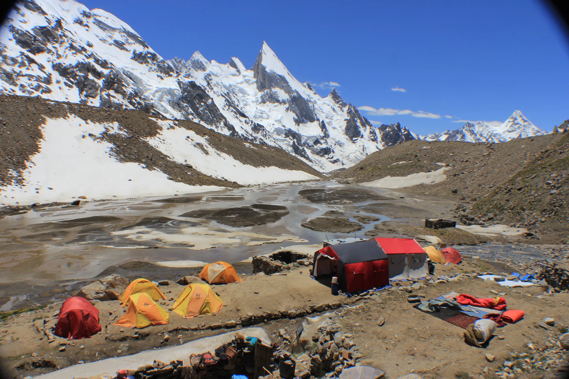 An expedition base camp with colorful tents sits beside a muddy riverbed, with sharp, snow-capped mountain peaks rising under a clear blue sky in the background.
