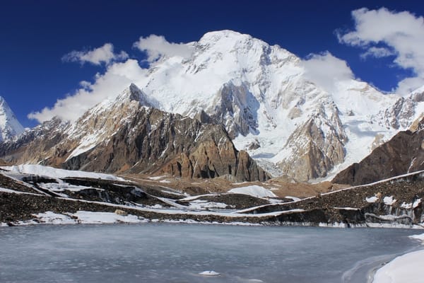 A frozen body of water reflects light in the foreground with a towering, snow-covered mountain massif rising dramatically under a deep blue, cloud-dotted sky.
