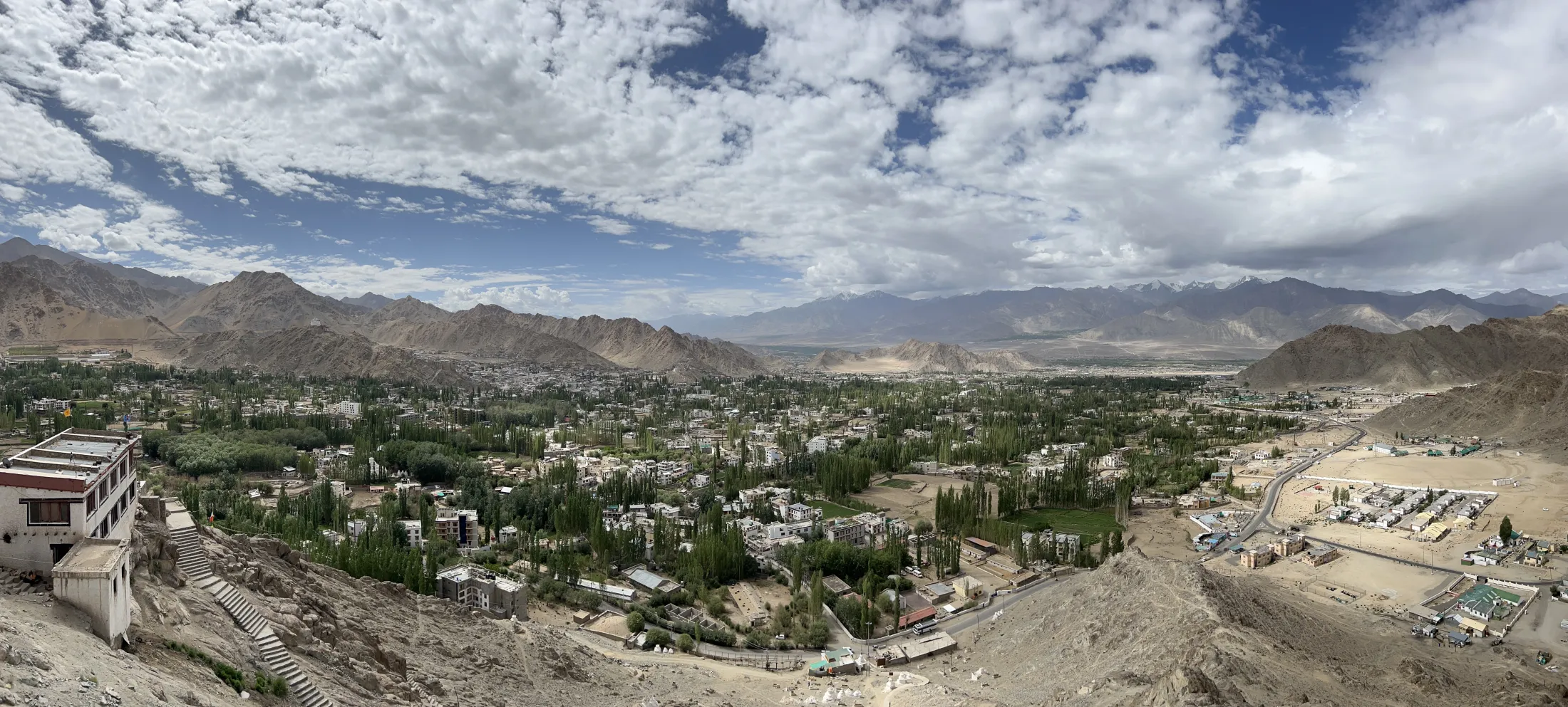 A wide panoramic view shows a green, tree-filled valley town nestled between barren, rocky mountain ranges under a sky filled with white clouds.
