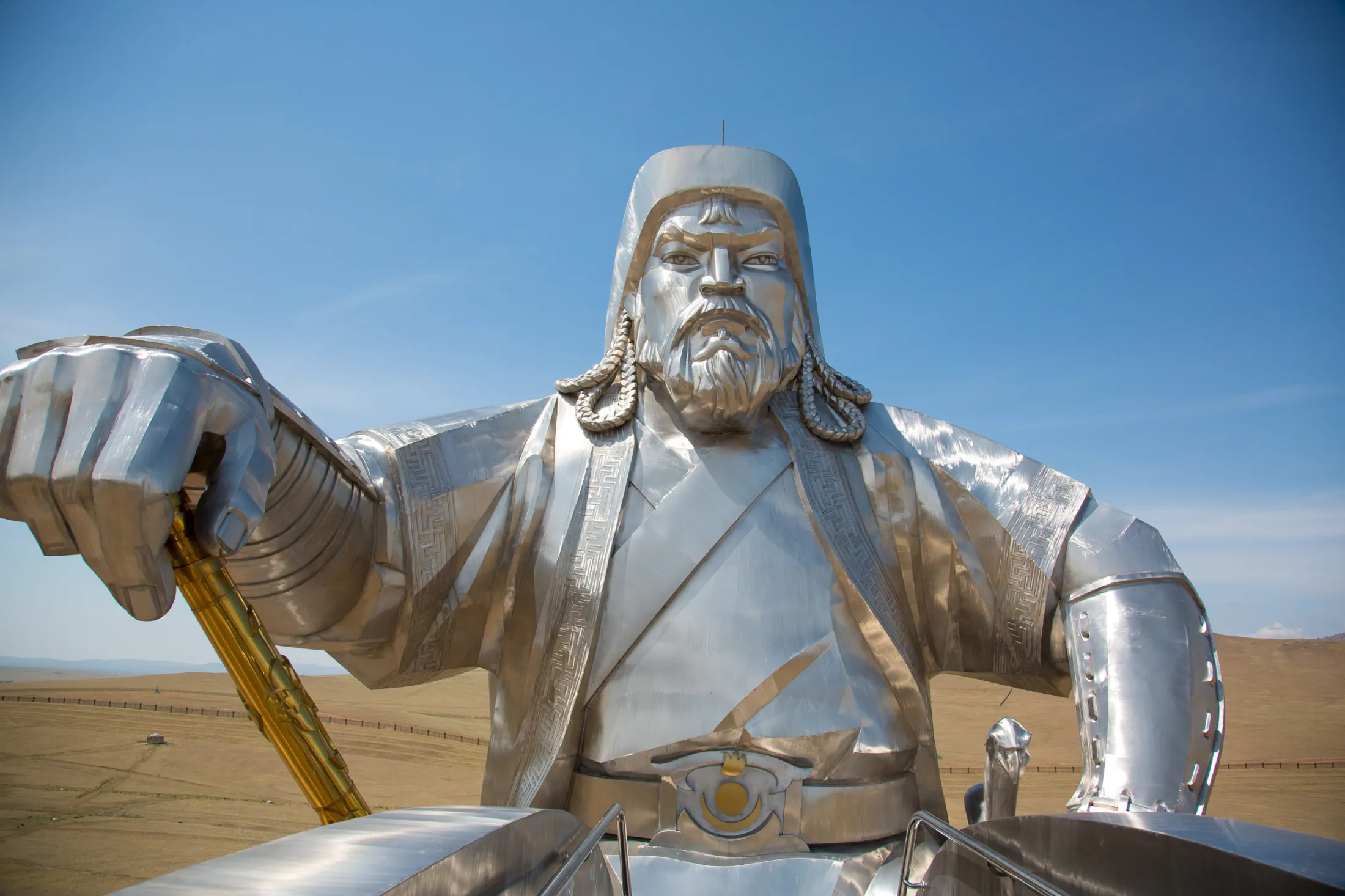 The colossal Genghis Khan Equestrian Statue made of gleaming stainless steel is depicted from a low angle against a clear blue sky and a barren plain.