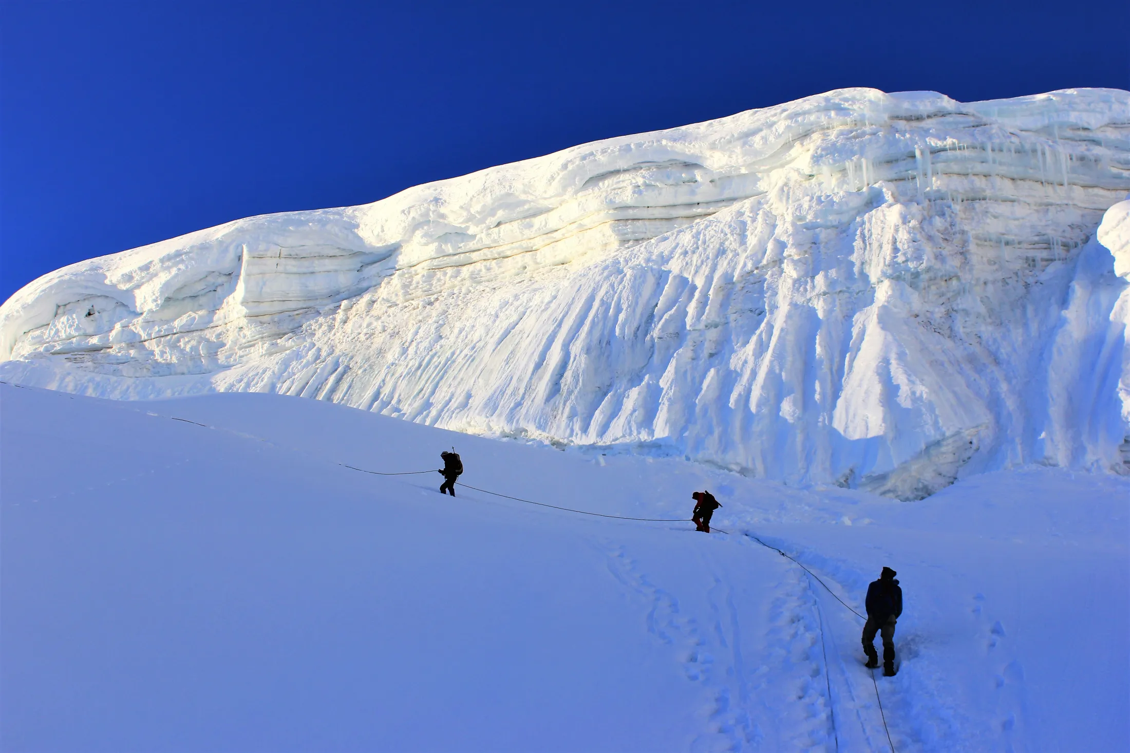 Climbers roped together ascend a steep, snow-covered slope beneath a massive, icy wall and serac formation under a clear blue sky.