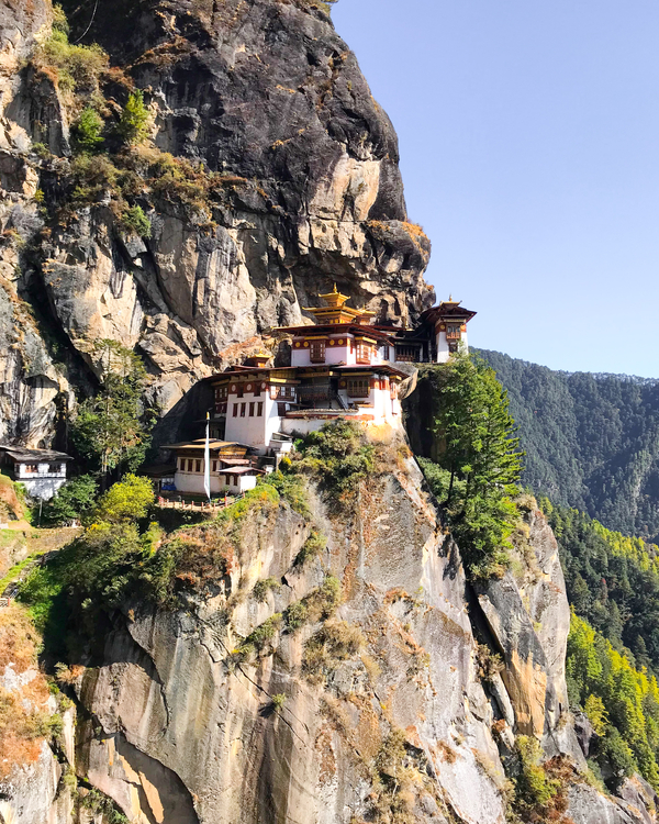Vertical view of the Tiger's Nest Monastery built onto a sheer cliff face surrounded by mountain slopes and a clear blue sky.