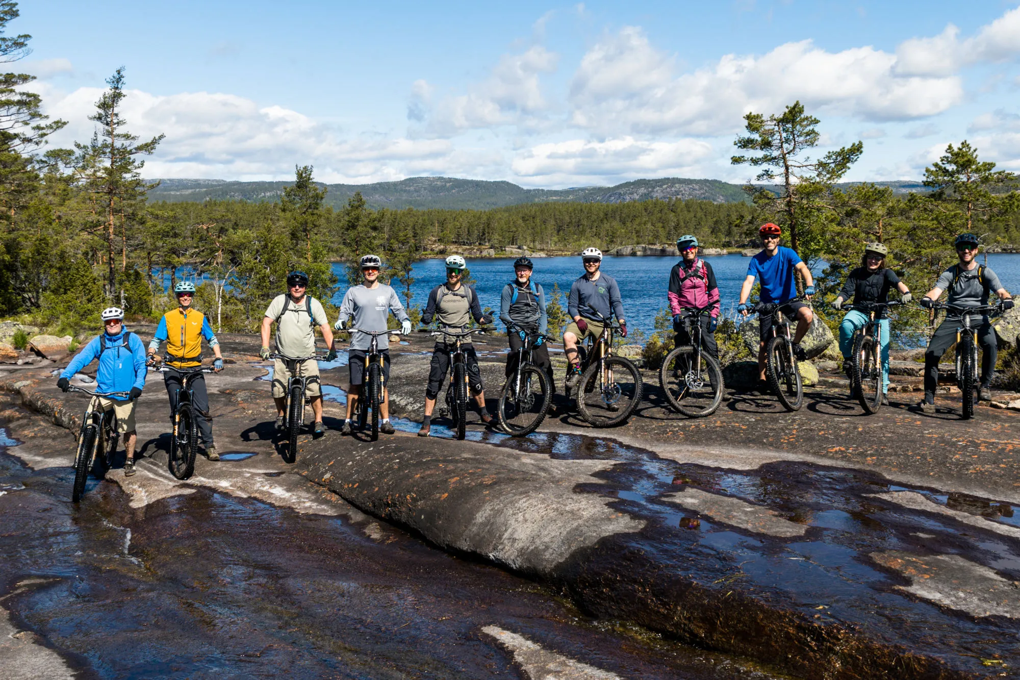 A group of mountain bikers posing on wet, smooth rocks by a lake with a forest in the background.