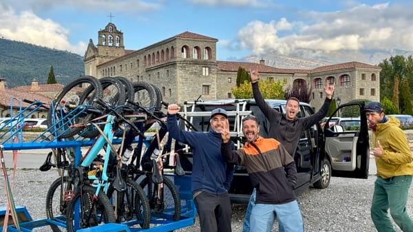 Four happy men posing with a bike trailer loaded with mountain bikes in front of a stone building.