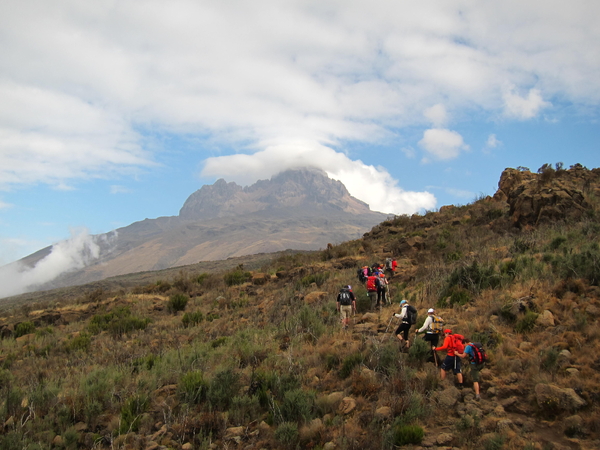 A group of hikers ascends a grassy mountain slope towards a towering peak shrouded in clouds.