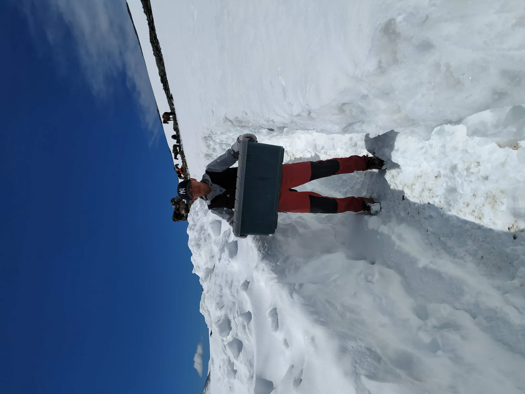 A person wearing a baseball cap and outdoor gear stands in a deep trench cut through the snow, holding a large grey plastic storage box, with pack animals visible on the ridge behind them under a blue sky.