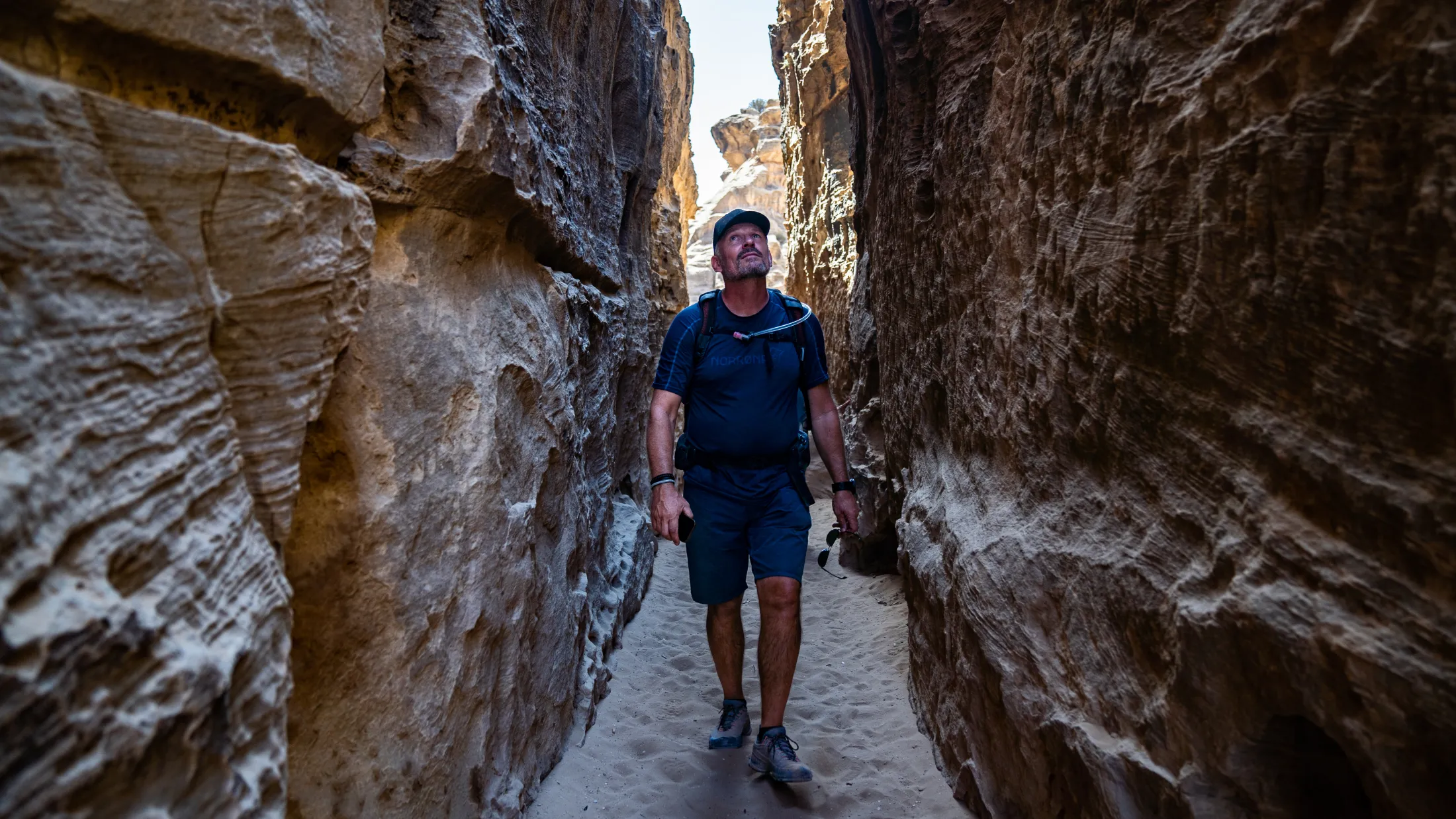 A man carrying a backpack and wearing a cap walks through a narrow canyon between tall sandstone walls, looking up toward the light.