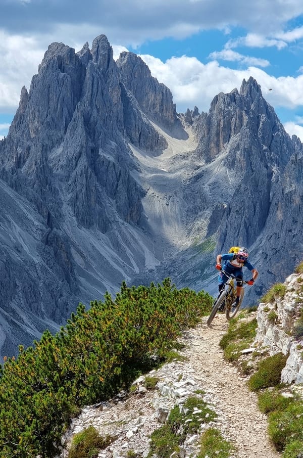 A mountain biker rides along a narrow alpine trail with towering, jagged peaks in the background.