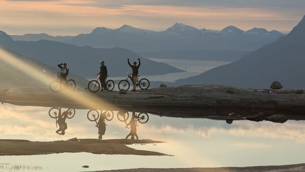 Three mountain bikers silhouetted against a mountain range and reflecting in a body of water.